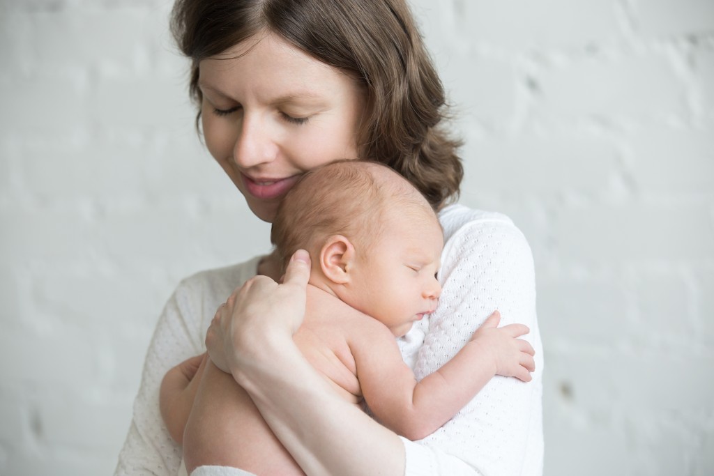 Portrait of young happy mother tenderly hugging sleeping adorable new born child and smiling. Funny newborn babe napping in mom arms. Mum and healthy little kid. Love, bonding, happy family concept.